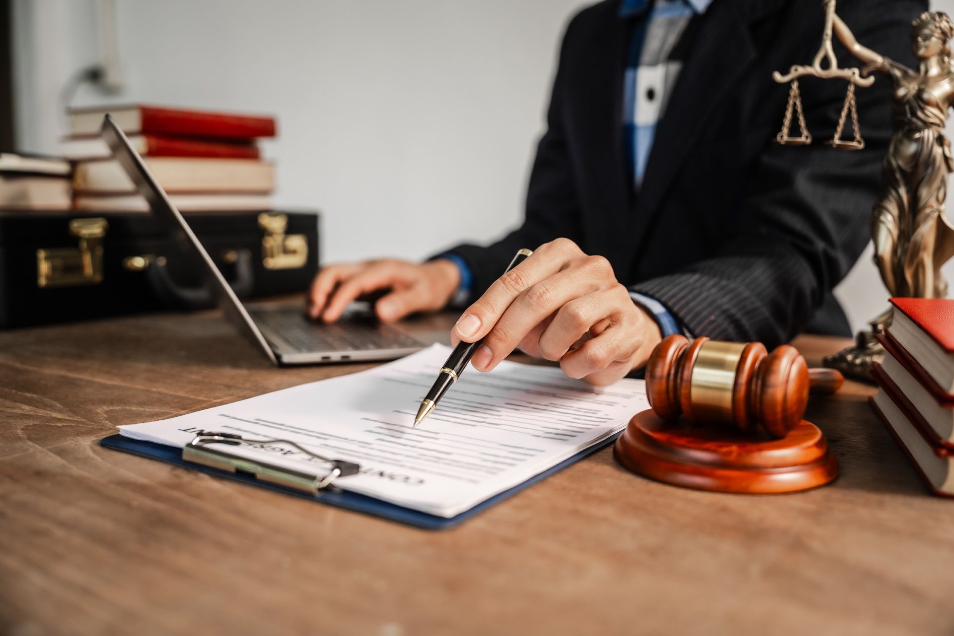 Lawyer at desk points to a document with pen, scales of justice, gavel, books, and laptop.