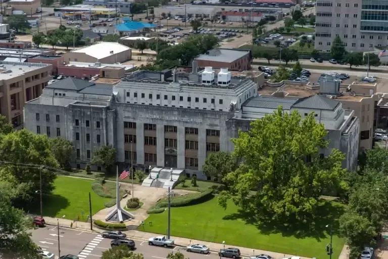 Aerial view of a large, stone courthouse with a green lawn and surrounding buildings in a city.