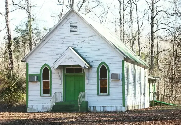 White church with green trim and door in a wooded area.