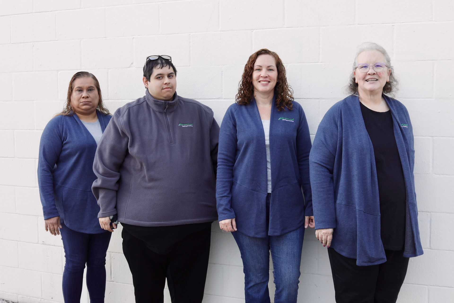 Four people standing in a row against a white brick wall, wearing casual blue cardigans or pullovers and smiling.