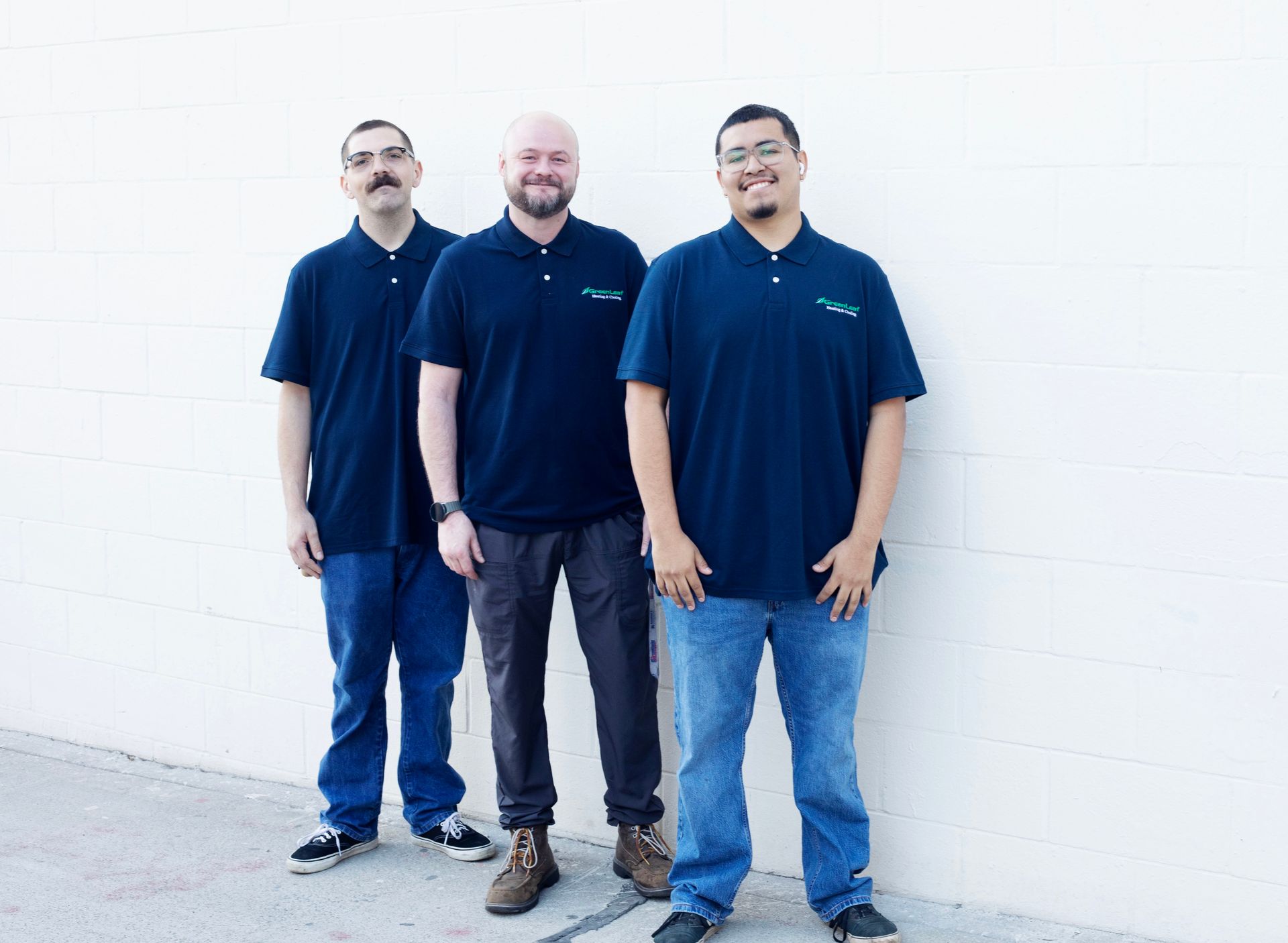 Three smiling individuals wearing matching navy polo shirts and jeans stand side-by-side against a plain white wall.