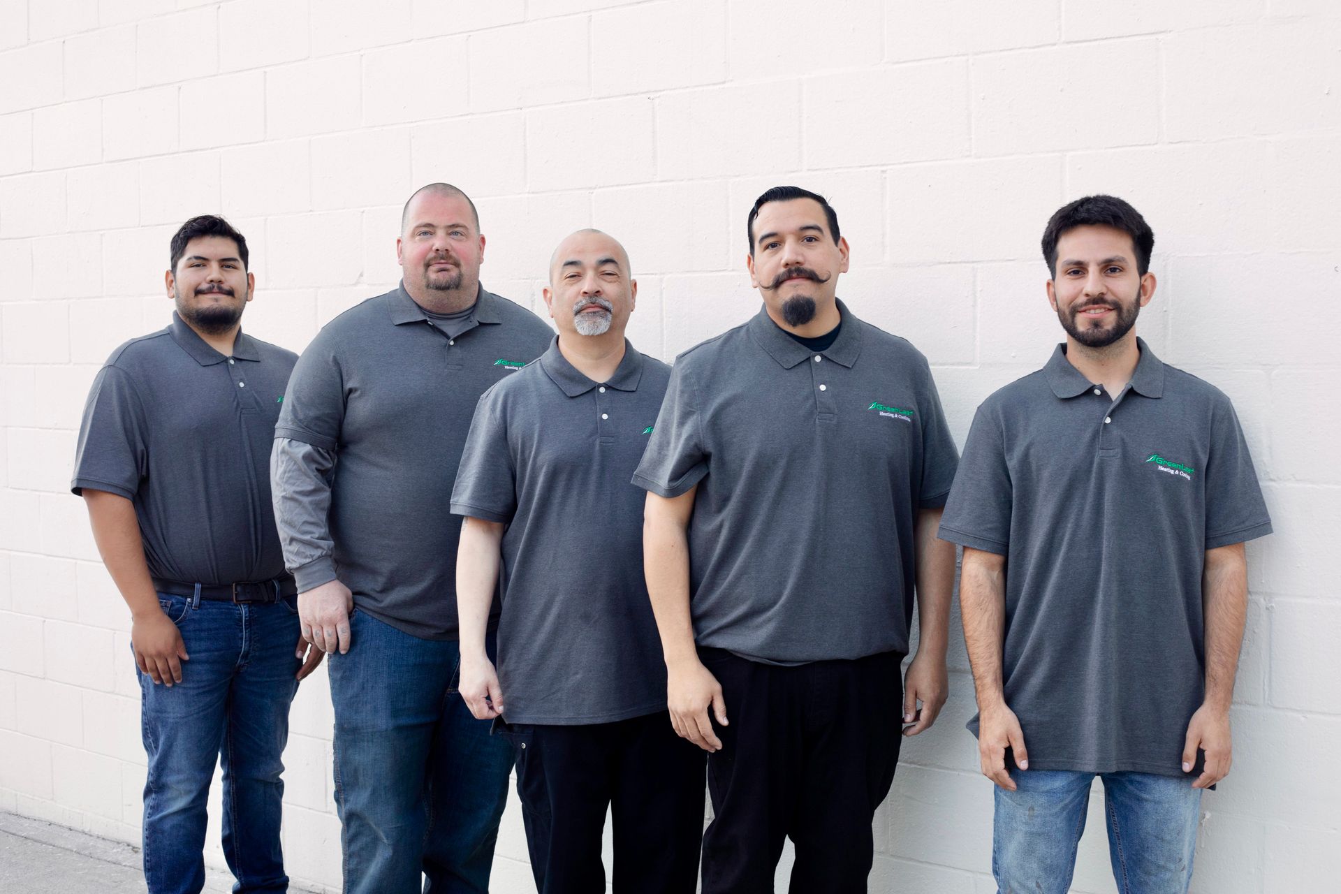 Five people wearing grey collared shirts stand in a row against a plain white wall.
