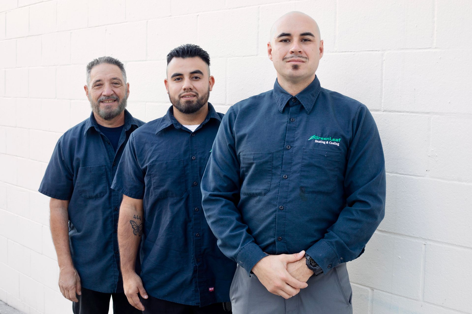 Three people in matching dark blue collared work shirts standing in a row against a white cinder block wall.