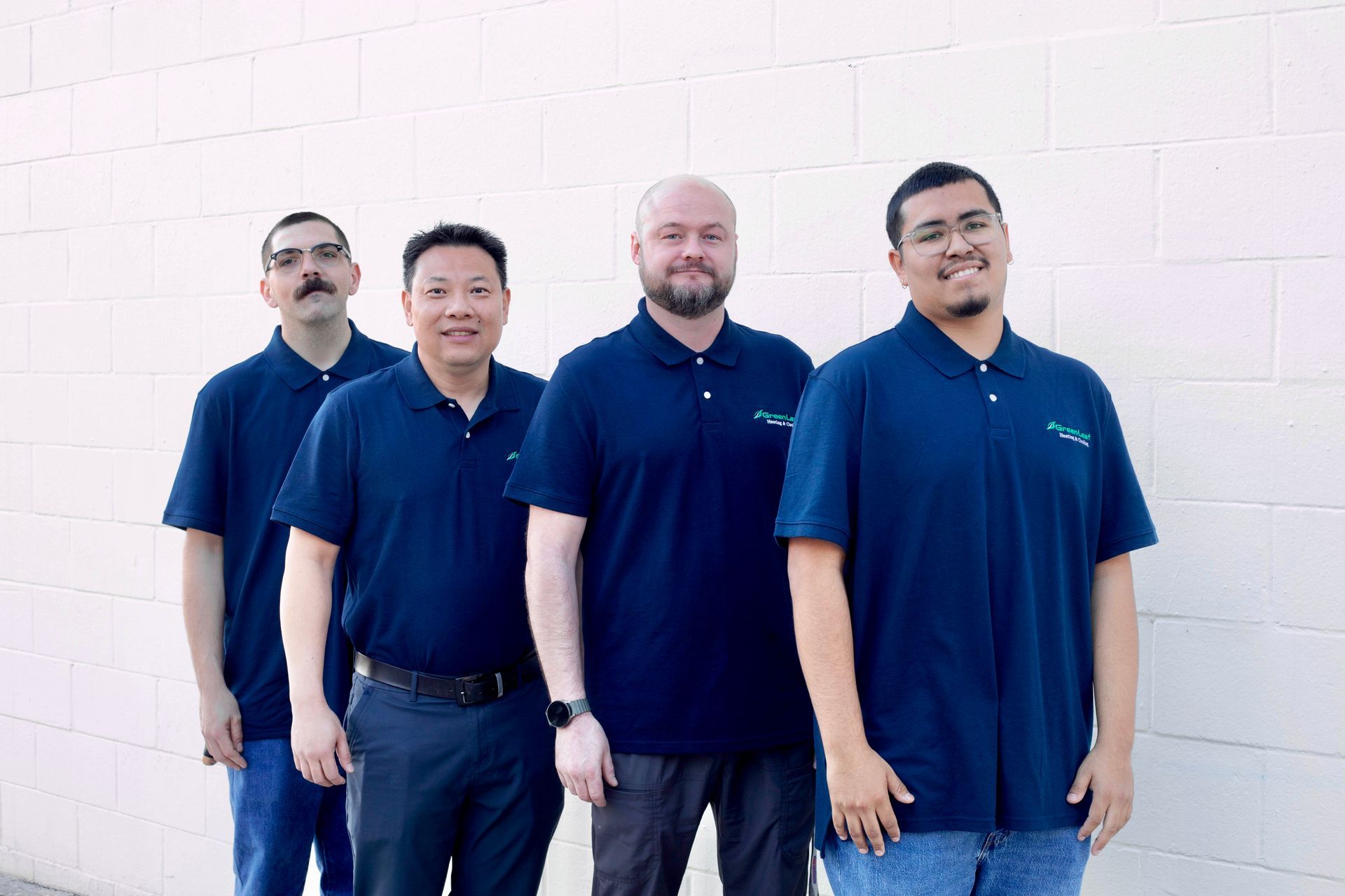 Four men stand side-by-side in matching navy blue polo shirts against a plain white brick wall.