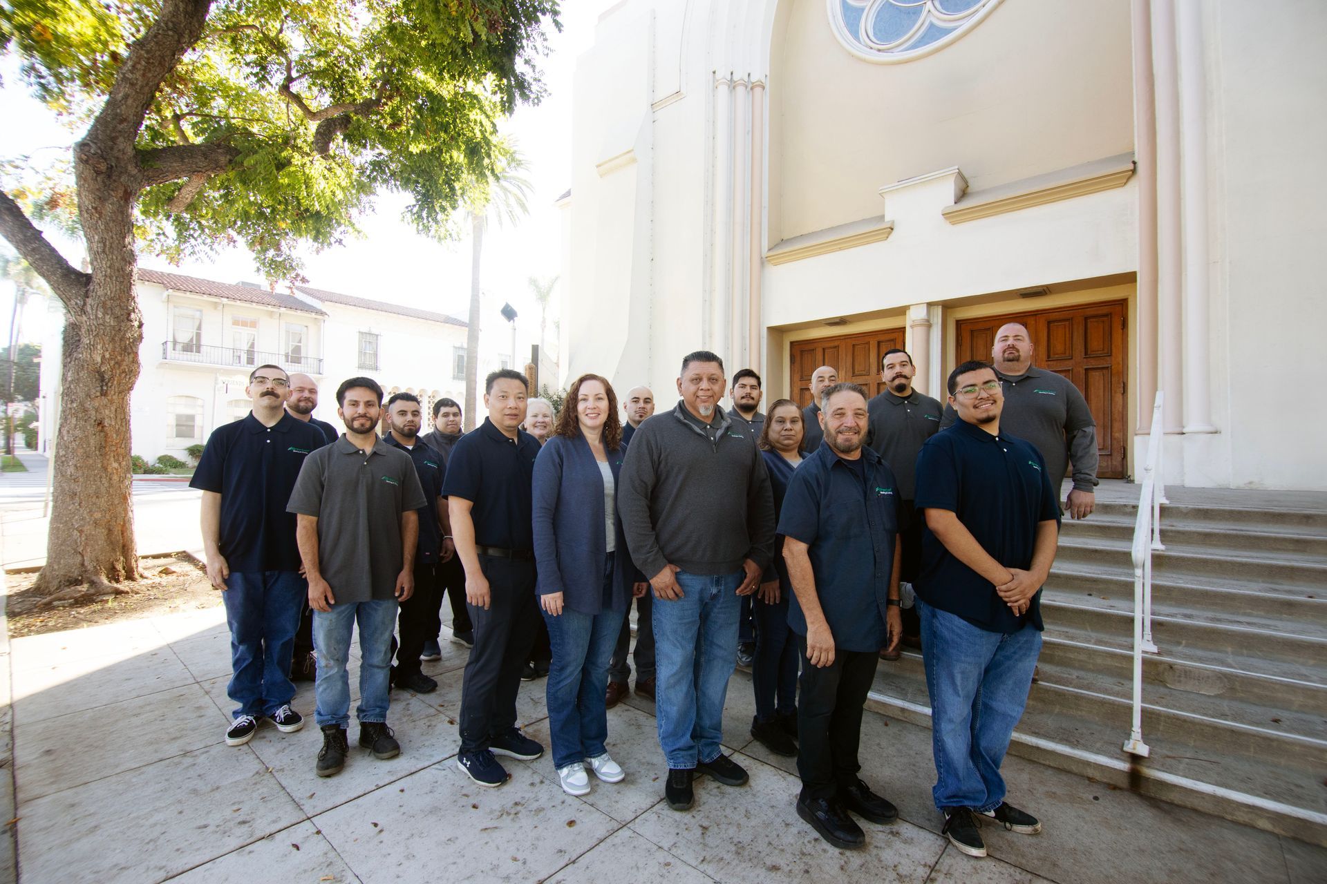 A group of people standing on the steps of a building, posed for a photo outside a church under a large tree.