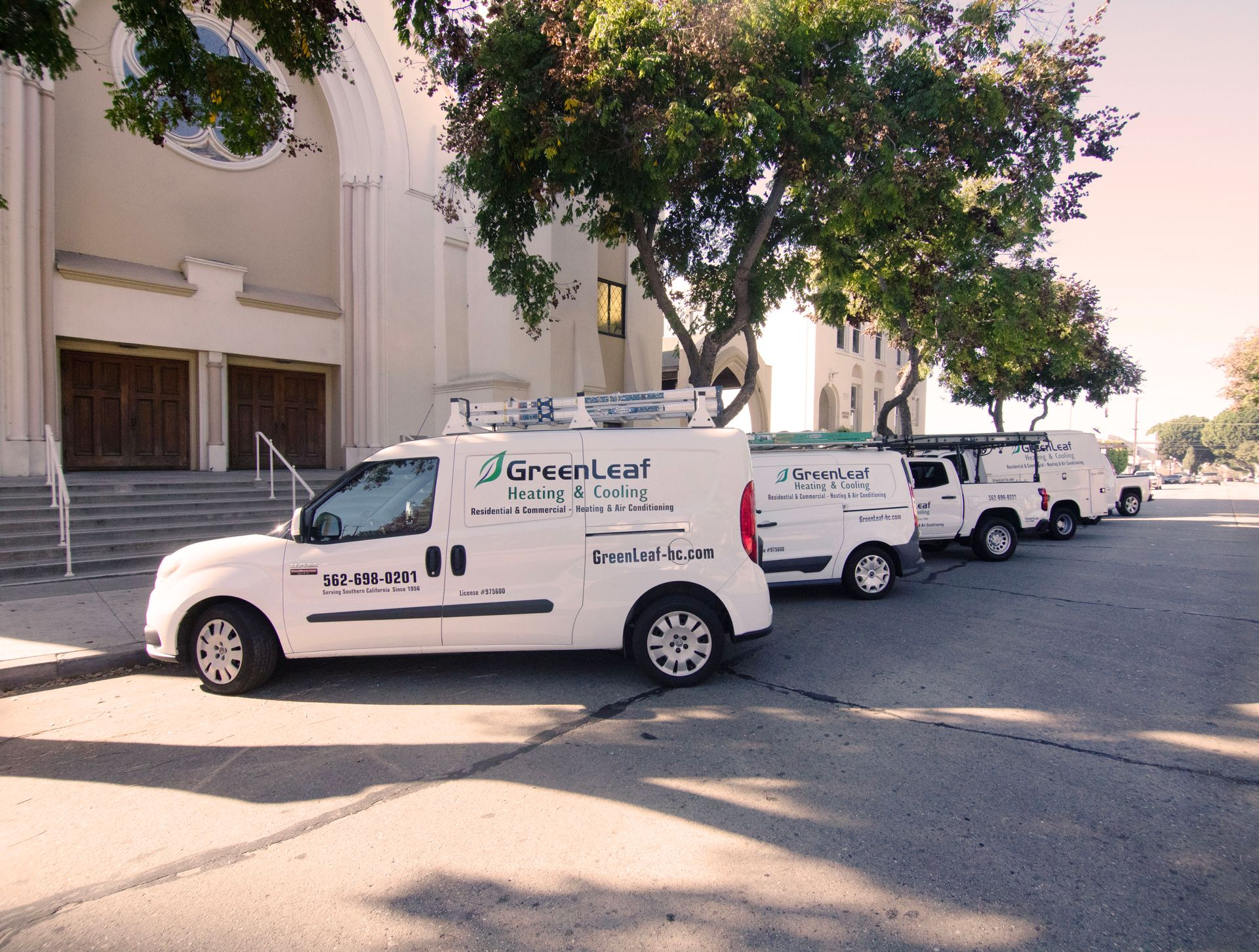 A line of white GreenLeaf Heating & Cooling service vans parked on a street in front of a large, light-colored building.