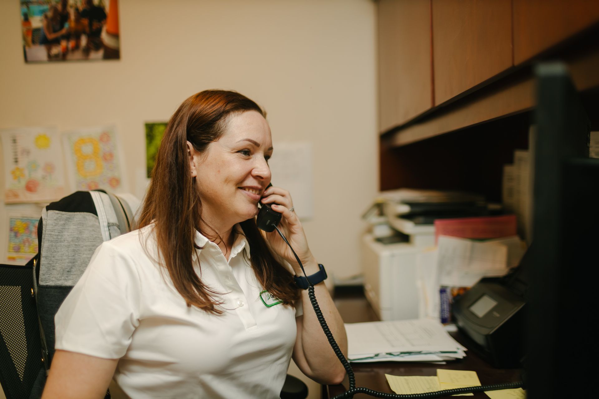 A person in a white polo shirt smiles while talking on a landline phone at a cluttered office desk.