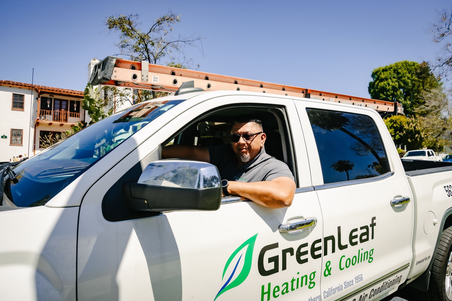 A smiling technician in a GreenLeaf Heating & Cooling service truck with a ladder on the roof.