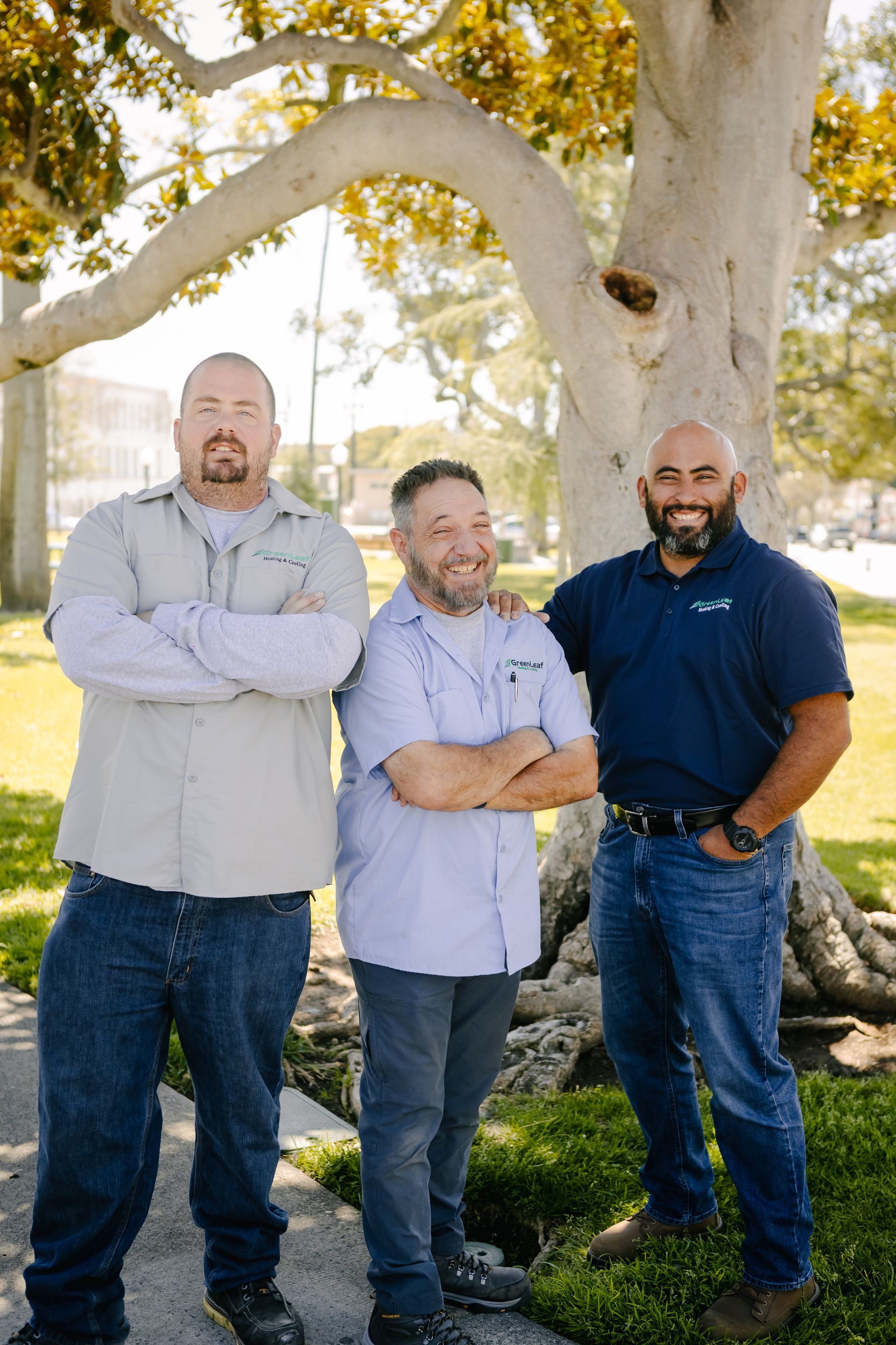 Three men stand side-by-side outdoors under a large tree, smiling and posing for a group portrait.