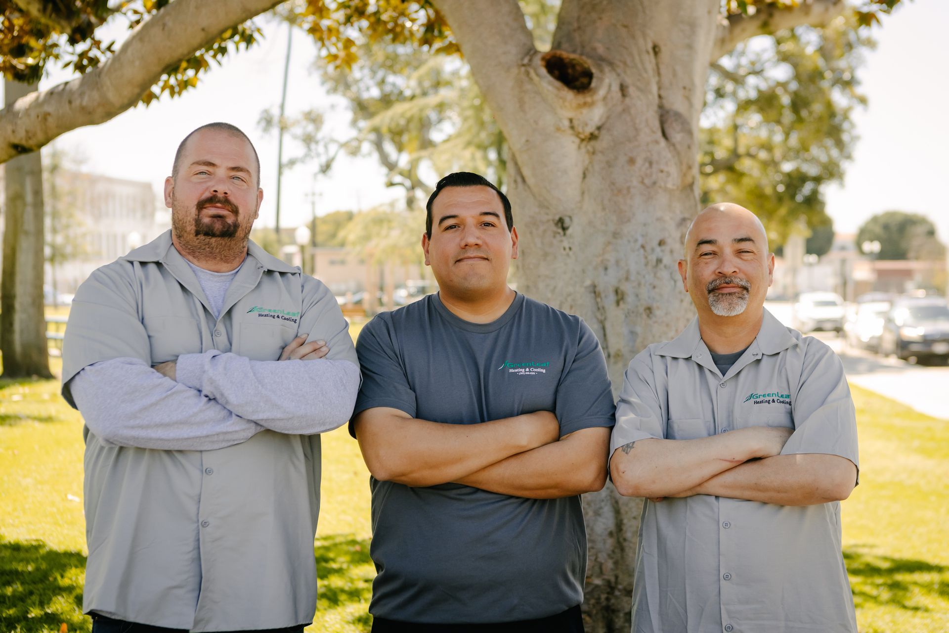 Three people with crossed arms standing side-by-side outdoors under a tree, wearing matching grey uniform shirts.
