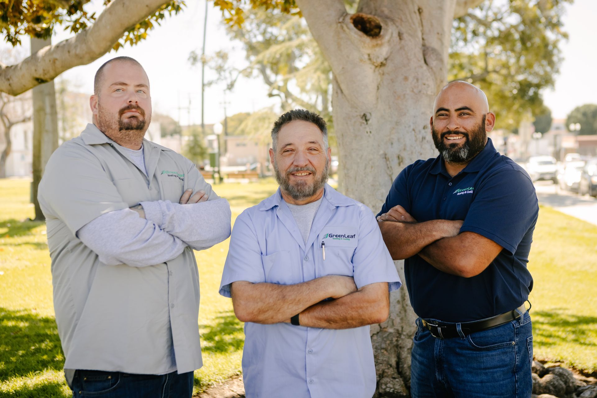 Three men standing with their arms crossed in front of a tree in an outdoor park setting.