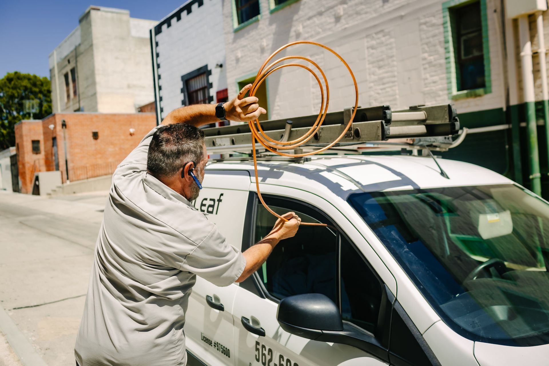 A person in work attire stands by a white service van, holding a coil of copper tubing near the open window.