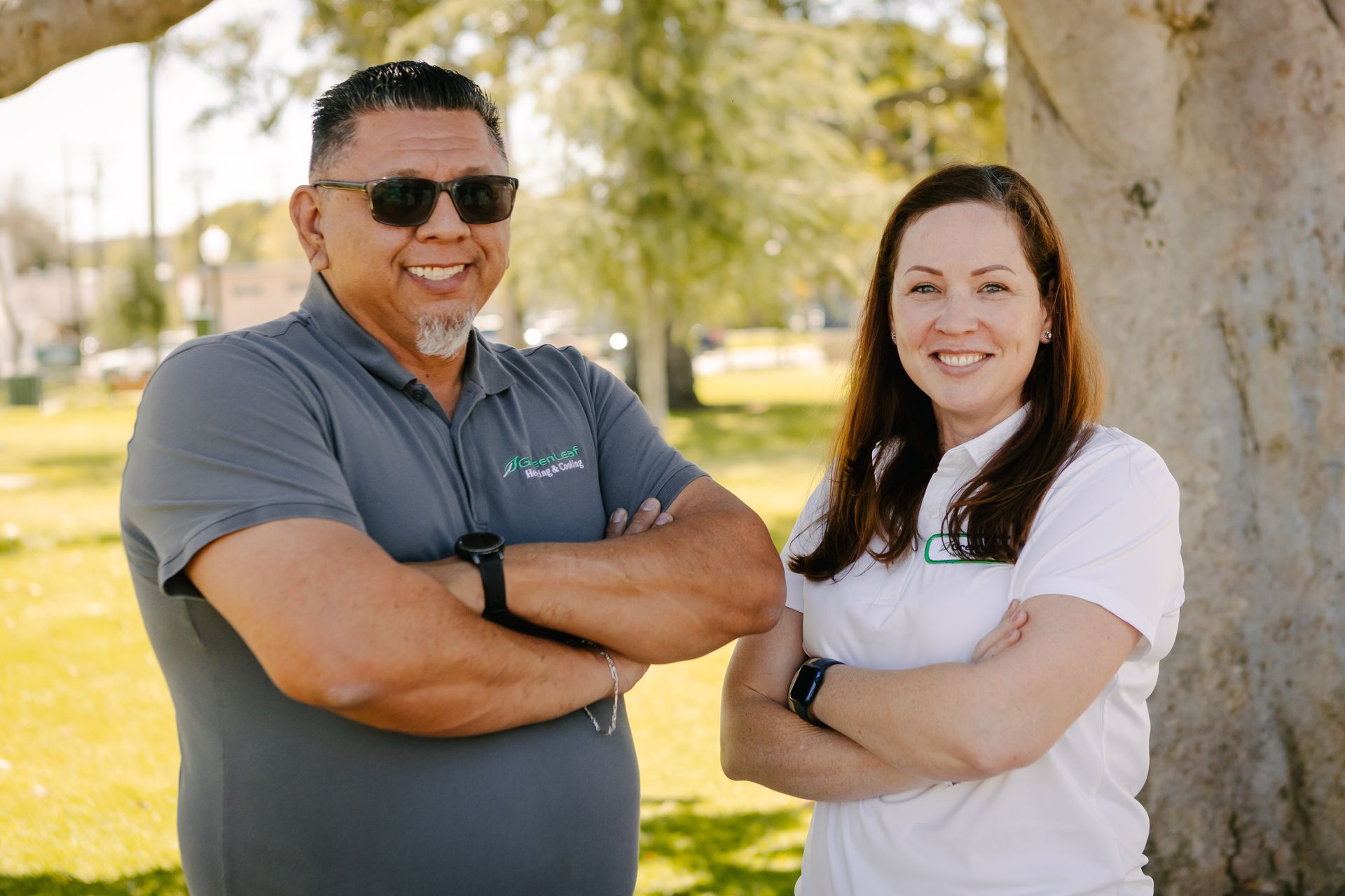 A man in a grey polo and a woman in a white polo stand with arms crossed in a sunny park with trees.