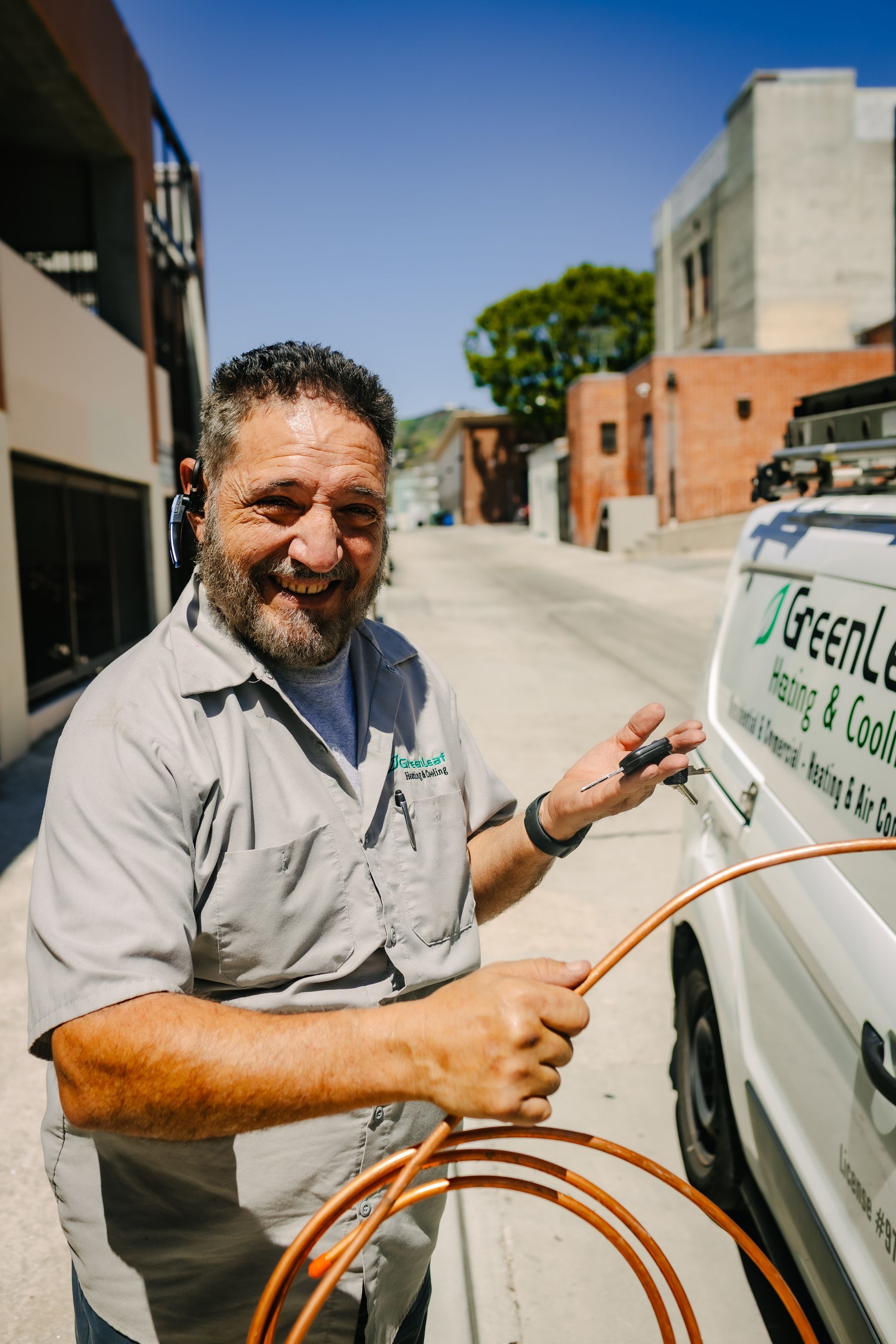 A smiling HVAC technician holding a copper pipe and keys next to a service van on a sunny street.