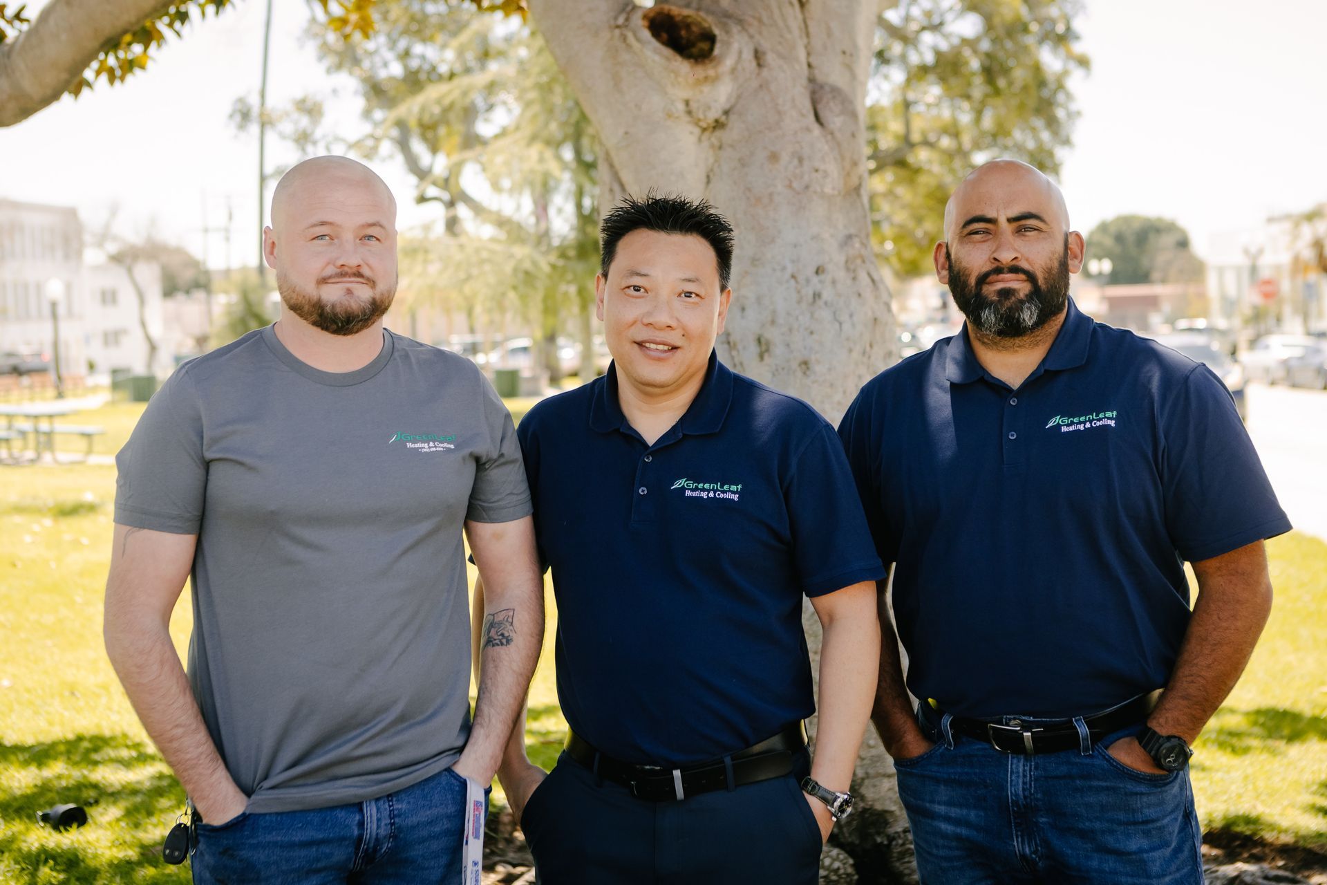 Three men stand side-by-side in front of a tree outdoors, wearing casual shirts and jeans, smiling toward the camera.