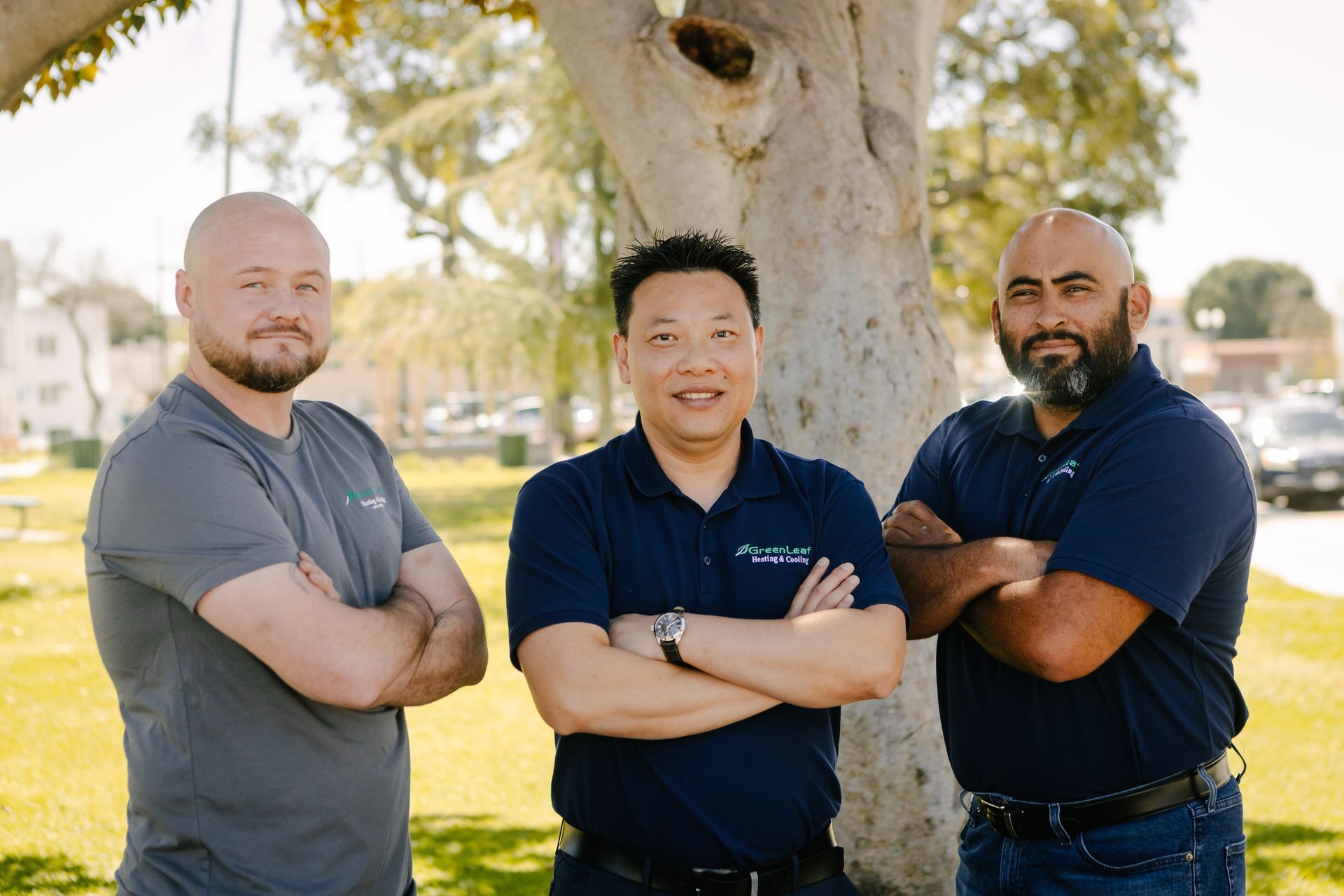Three people with crossed arms standing in a park in front of a tree.