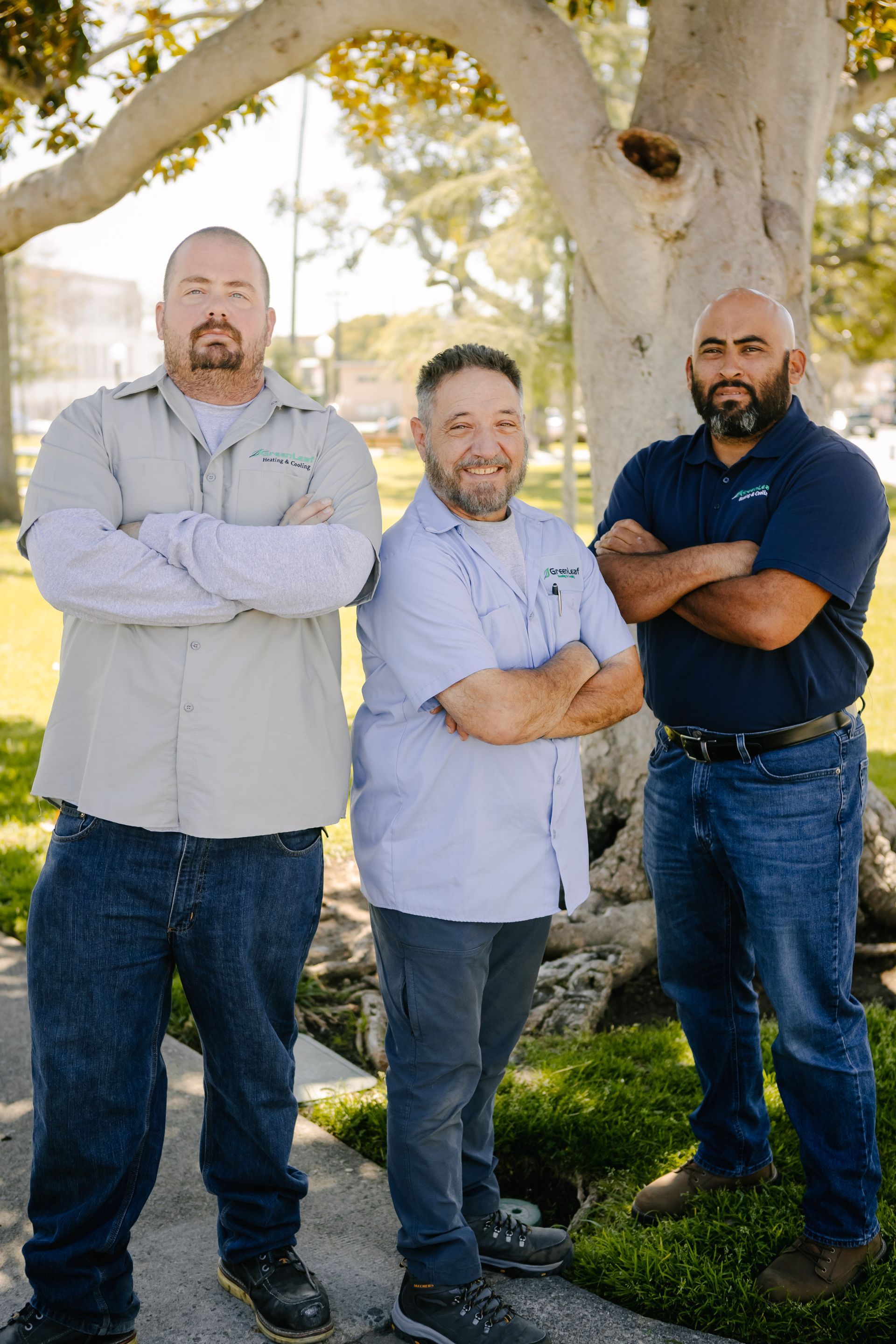 Three men stand with arms crossed before a large tree in an outdoor park setting.