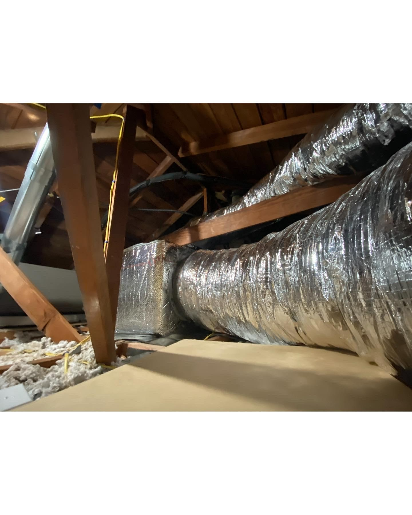 A metallic flexible HVAC duct connects to a rectangular vent box in a wooden attic space with loose-fill insulation.