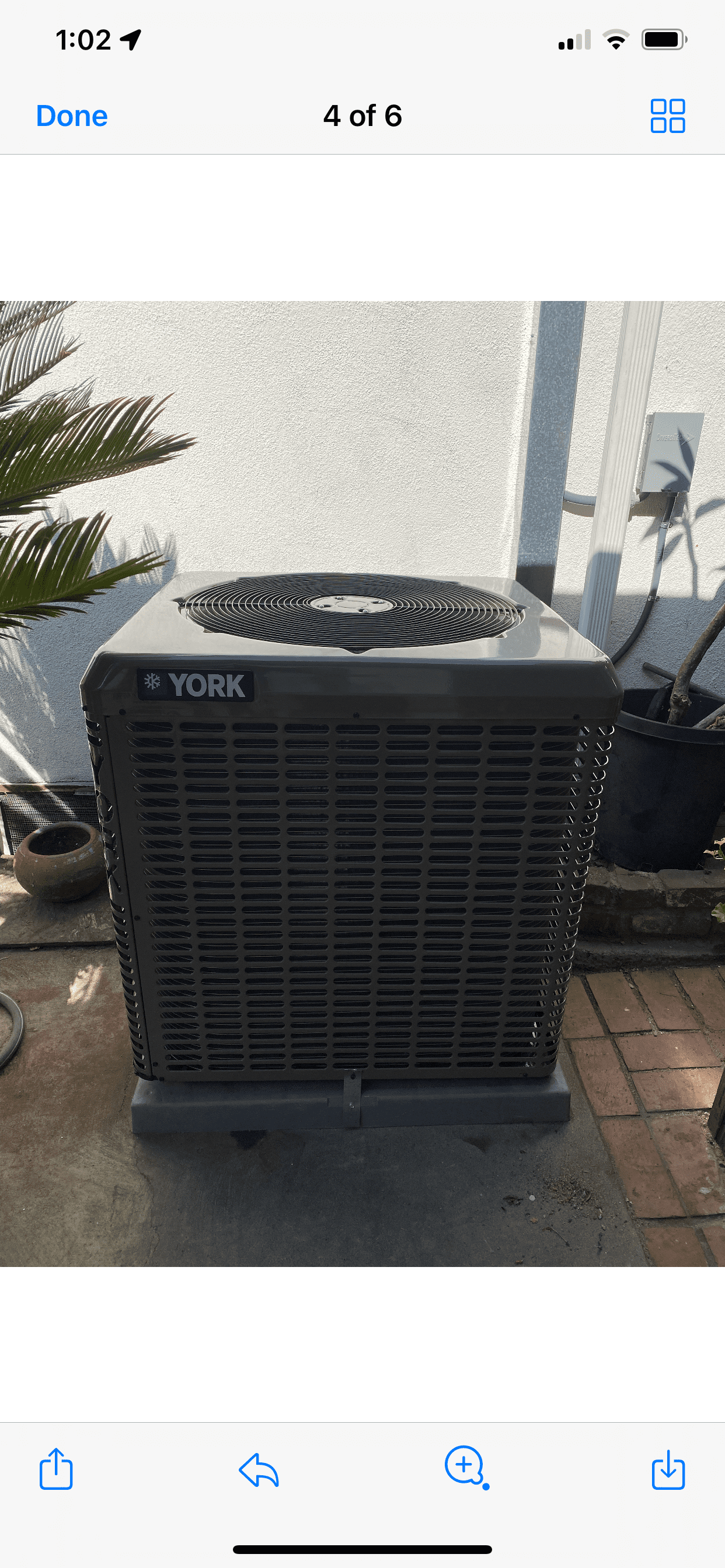 A dark gray outdoor air conditioning unit sits on a concrete patio next to potted plants.