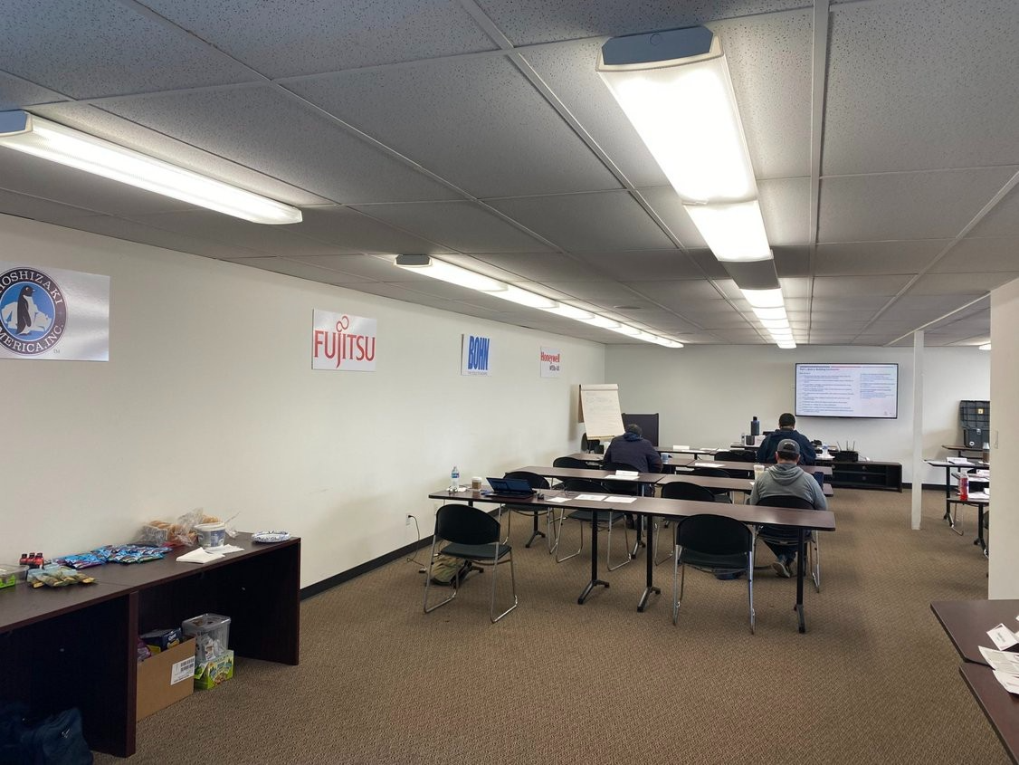 An office training room with rows of tables, chairs, and people working, featuring wall signage and overhead lighting.