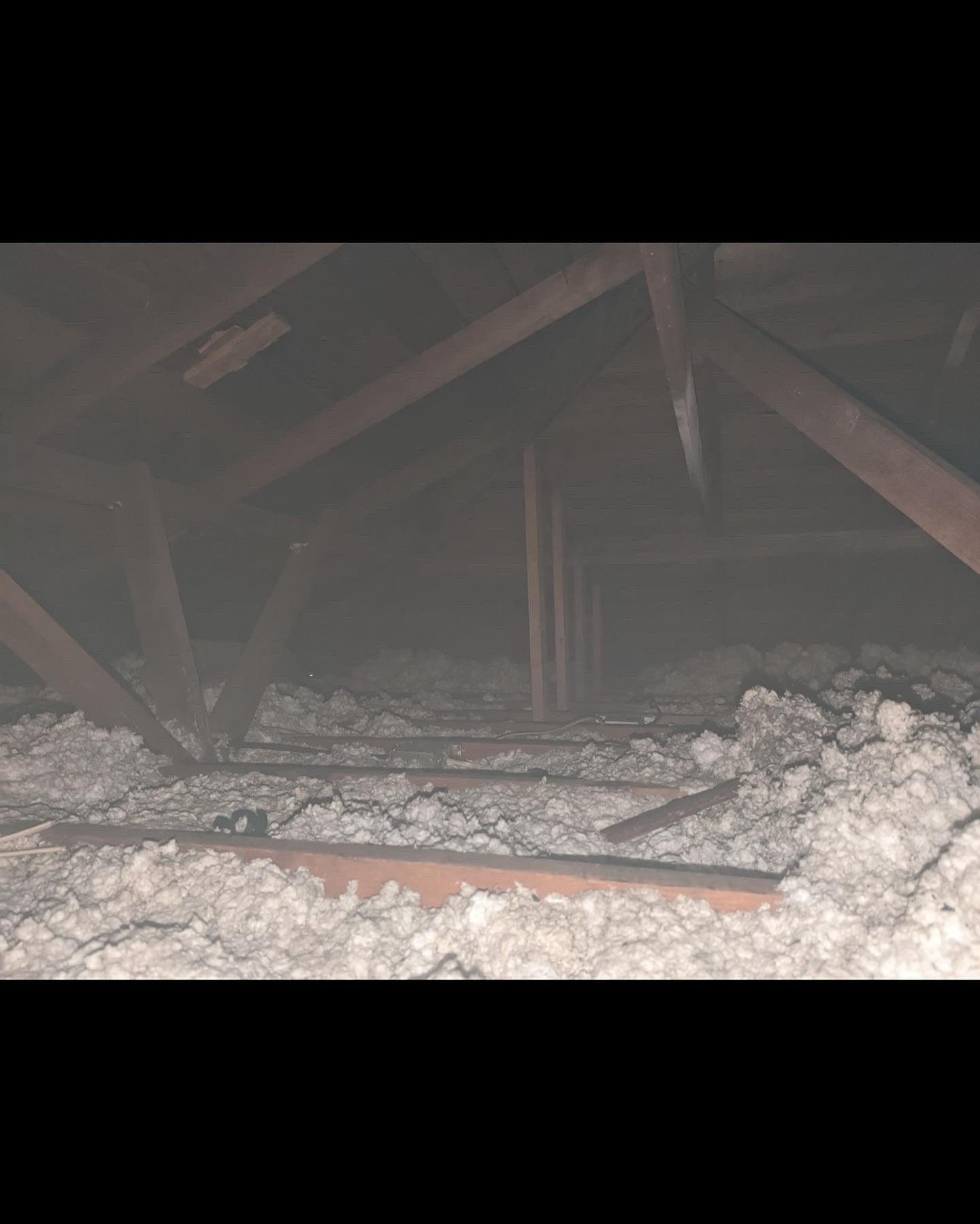 Dimly lit attic space showing wooden rafters, support beams, and loose-fill fiberglass insulation between floor joists.