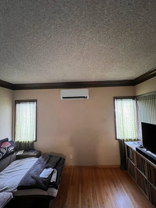 A living room with hardwood floors, a couch on the left, a TV console on the right, and a wall-mounted AC unit.