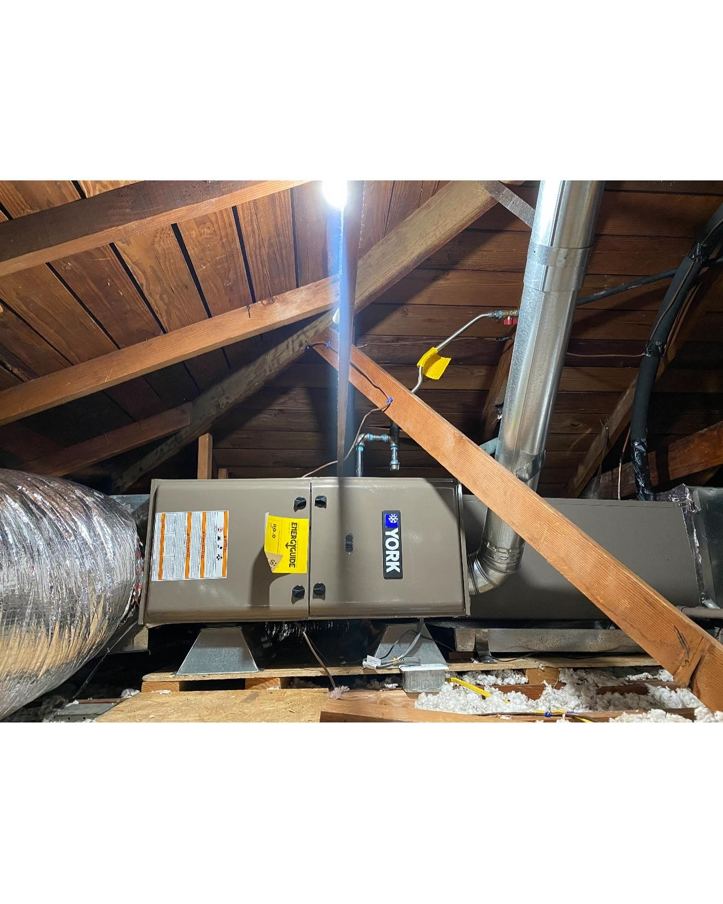 An HVAC air handler unit installed in an attic crawl space with wooden rafters, silver ducting, and a warning label.