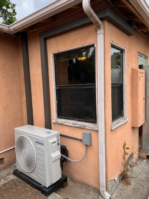An outdoor air conditioning unit sits on a black pad against the side of a tan stucco house next to a window.