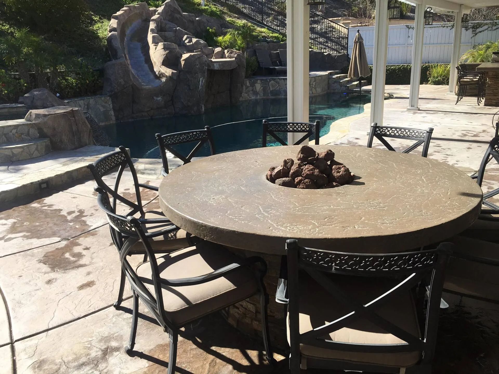 Round outdoor dining table with chairs, poolside, with water slide and rocks in background.