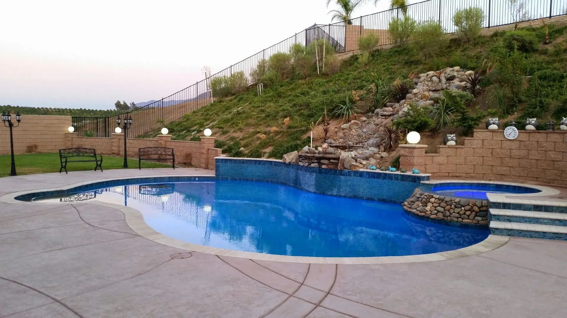 Pool with blue tiles and waterfall feature, beside a green hill.