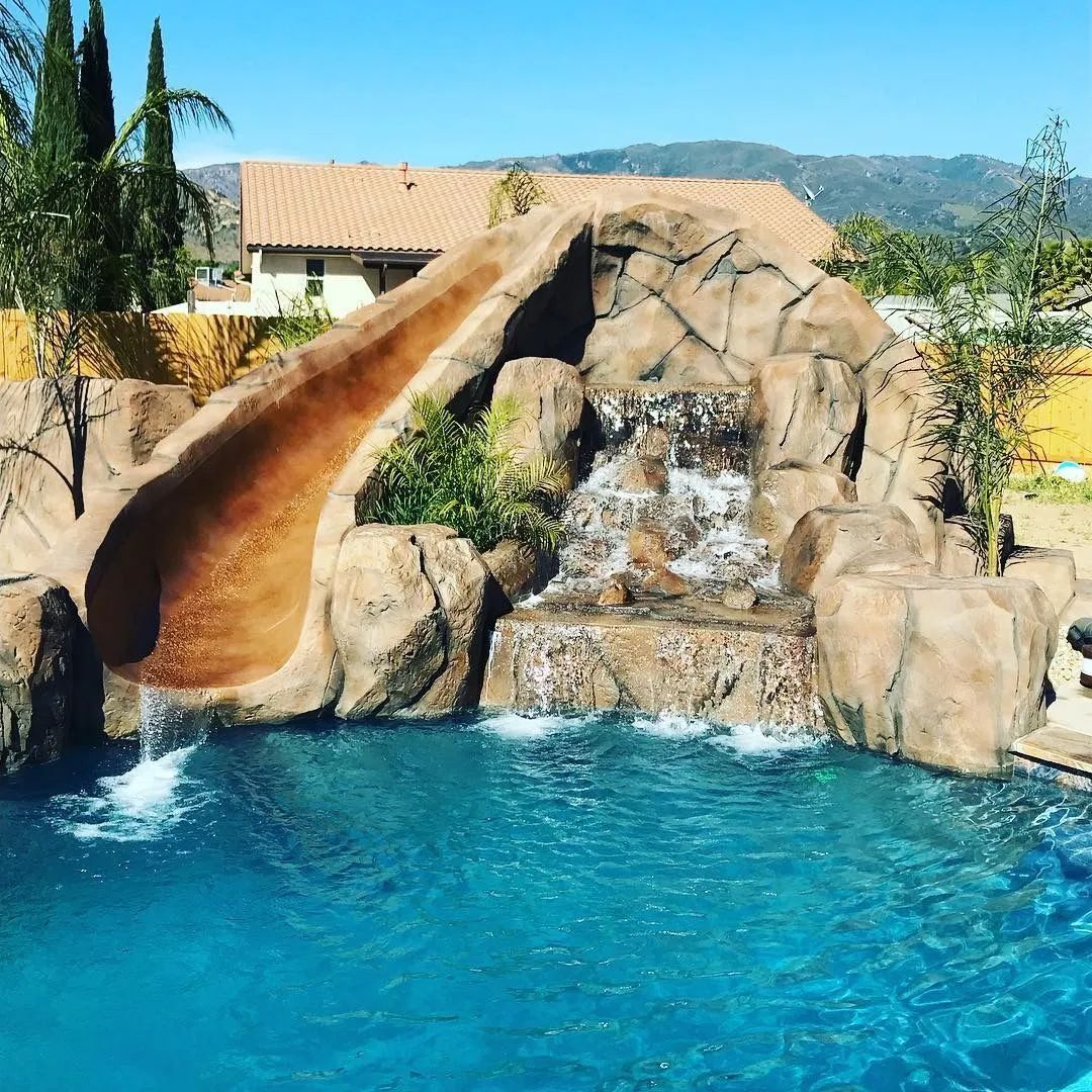 Pool with waterfall feature and water slide; mountains in the background, blue water, sunny day.