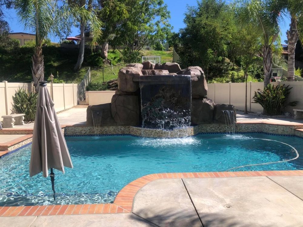 Pool with a rock waterfall feature, umbrella, and a tan fence in the background.