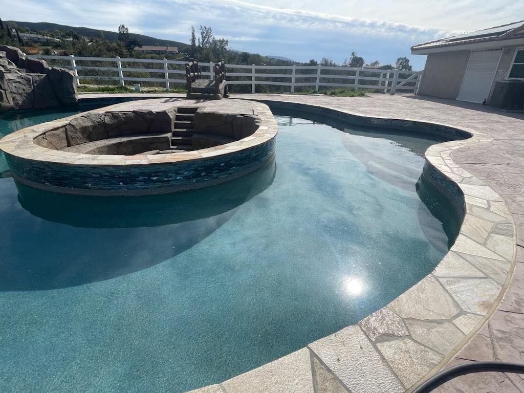 Pool with a circular hot tub, stone edging, white fence, and blue water on a sunny day.