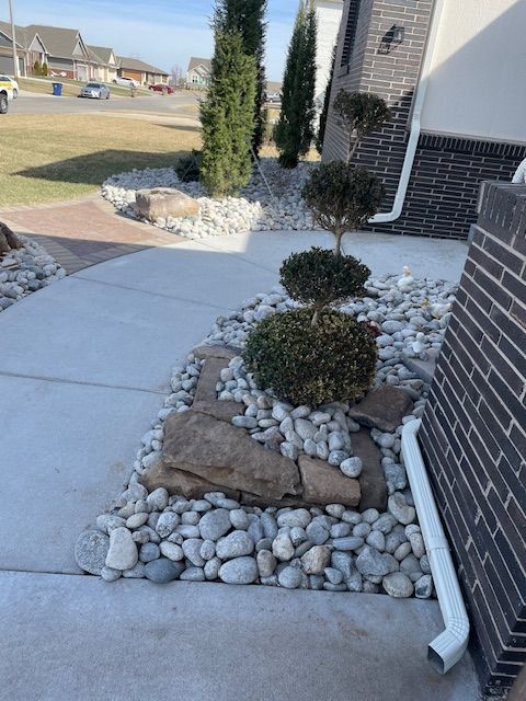 A decorative garden bed with rocks, a topiary shrub, and concrete pathway in front of a modern brick house.