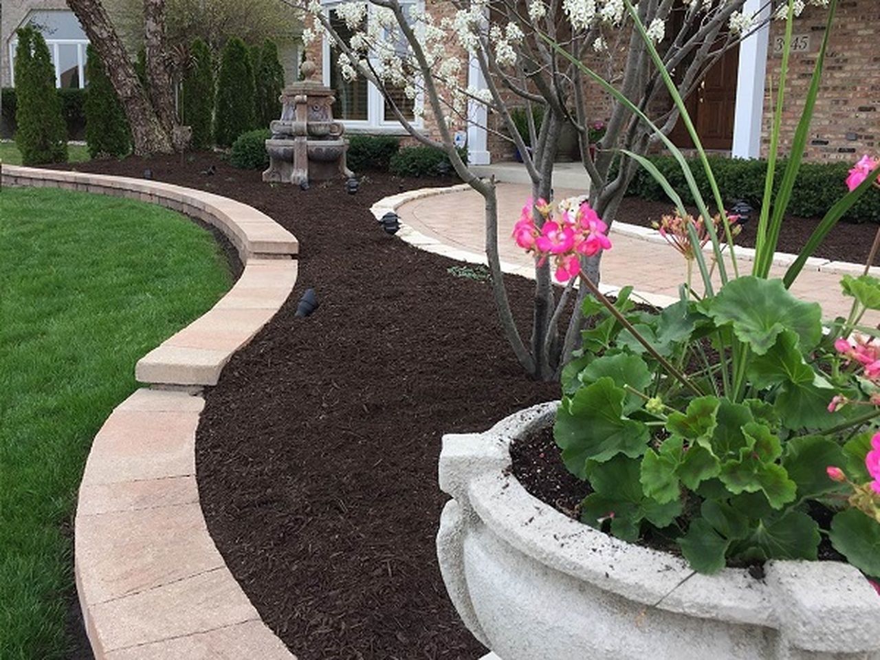 Curved walkway with stone border and mulch, leading to a house. Pink flowers in a stone planter.
