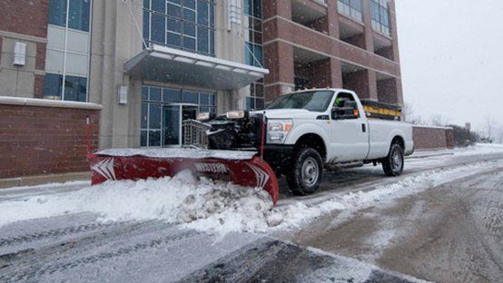 A white pickup truck with a red snowplow clears snow from a city street in front of a building.