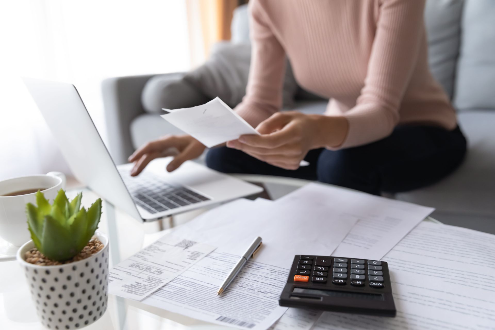 Person reviewing paperwork and calculator beside a laptop on a couch