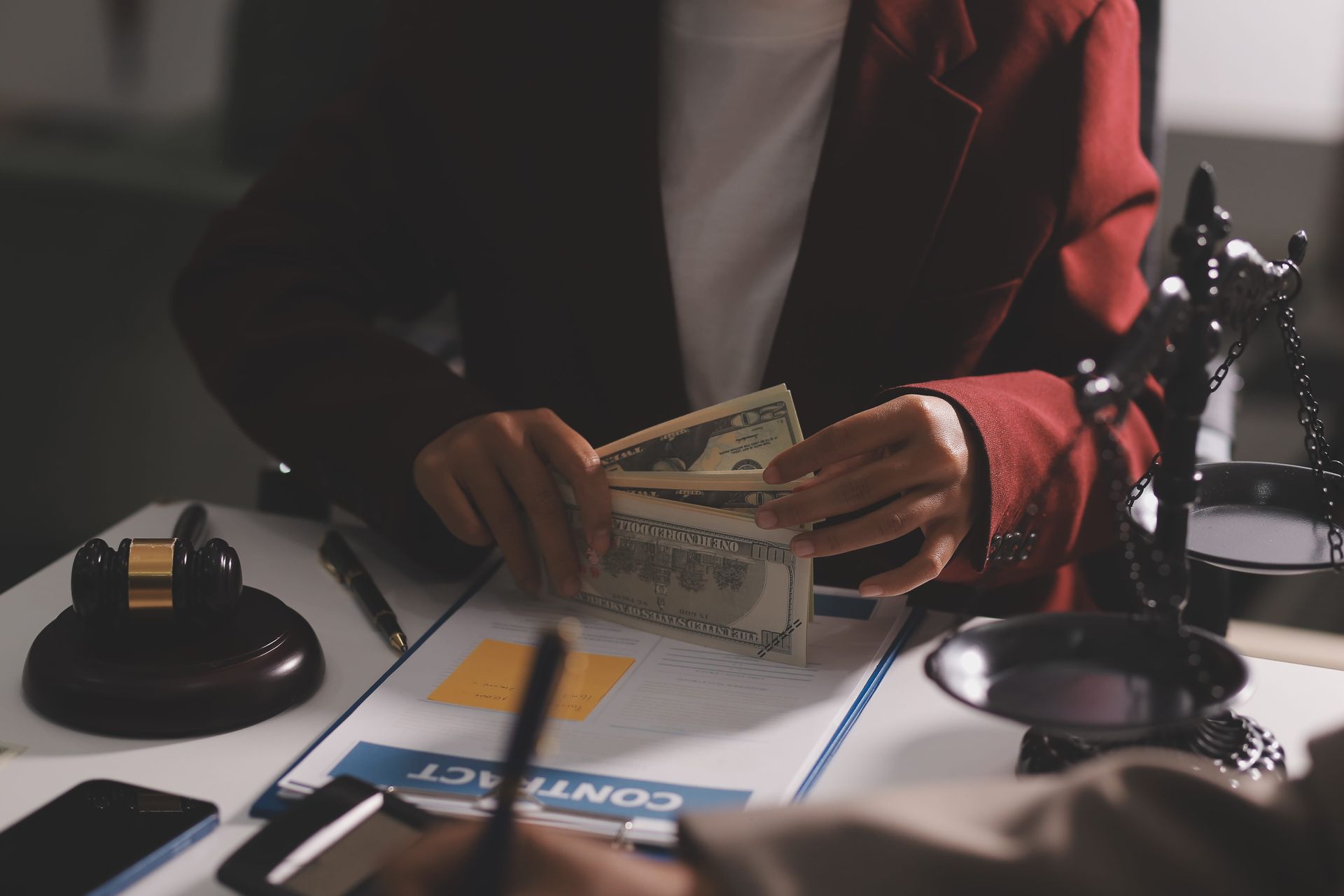 Person counting cash at a desk with a judge’s gavel, scales, and contract papers in view