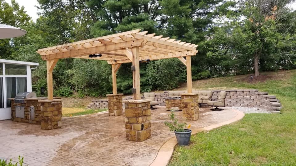 A wooden pergola with stone columns stands on a patio in a backyard, surrounded by greenery and a small retaining wall.