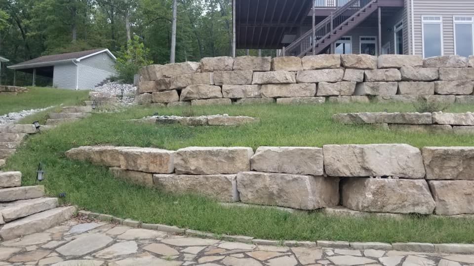 Stone retaining walls with grassy patches lead up to a house with a wooden deck, near a small building.