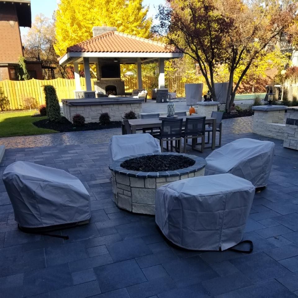 A patio with covered outdoor furniture and a fire pit. A gazebo sits in the background, beneath a tree with yellow leaves.