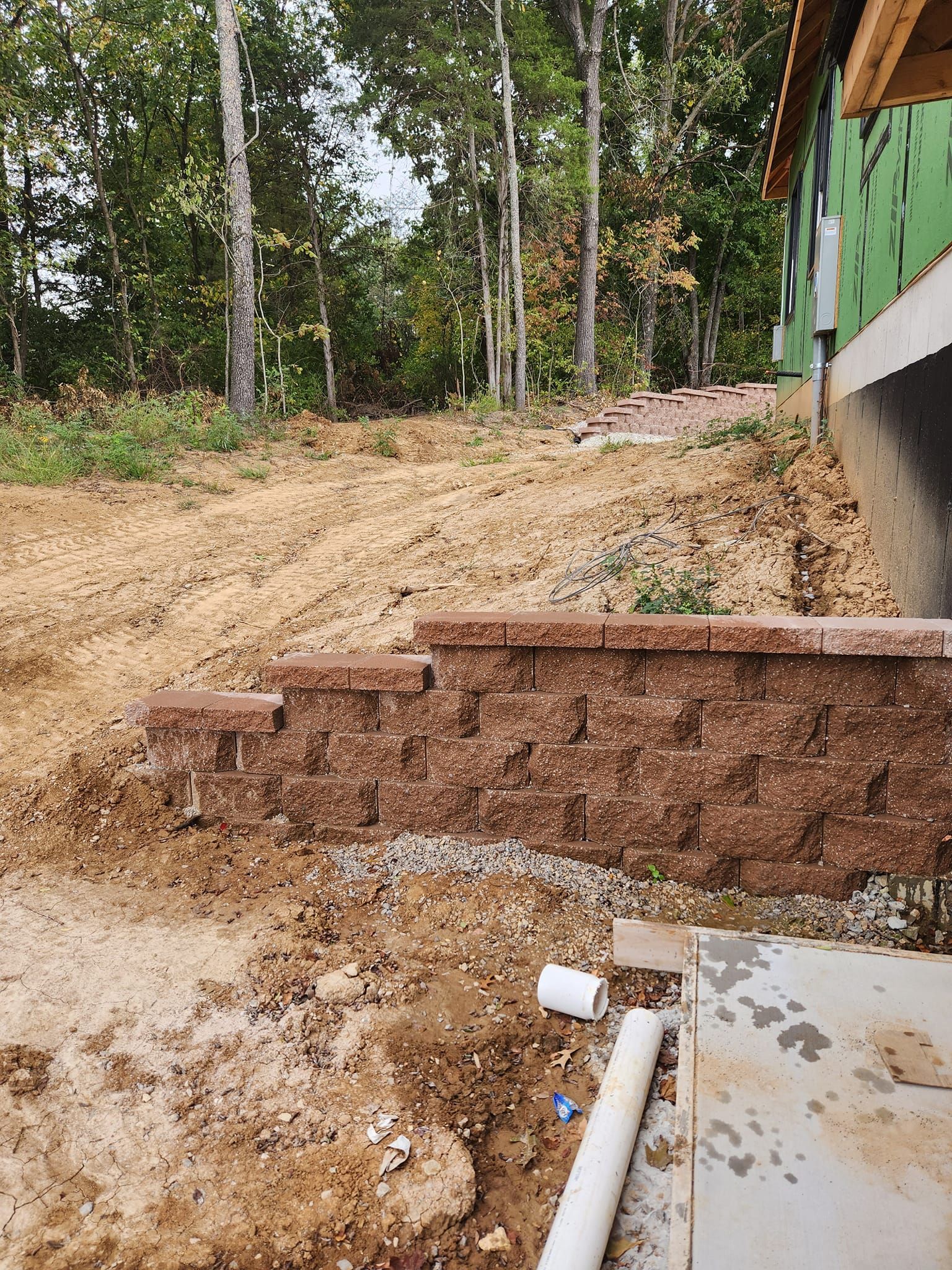 A brick retaining wall steps up a sloped, dirt yard near a partially constructed building. The background is a forest.