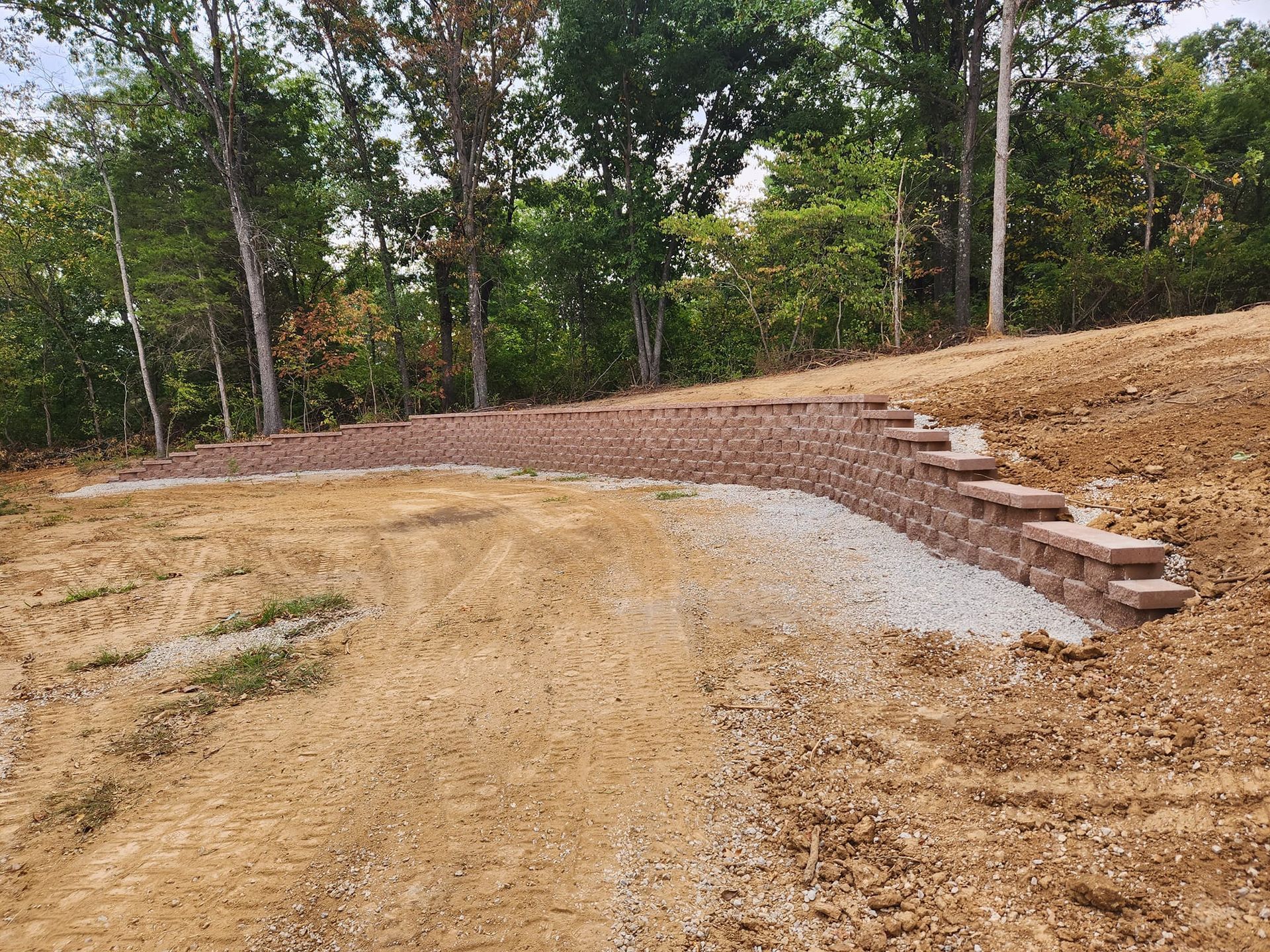 A terraced yard with retaining walls and stone steps leading up to a wooded area. The lower area is dirt and gravel.