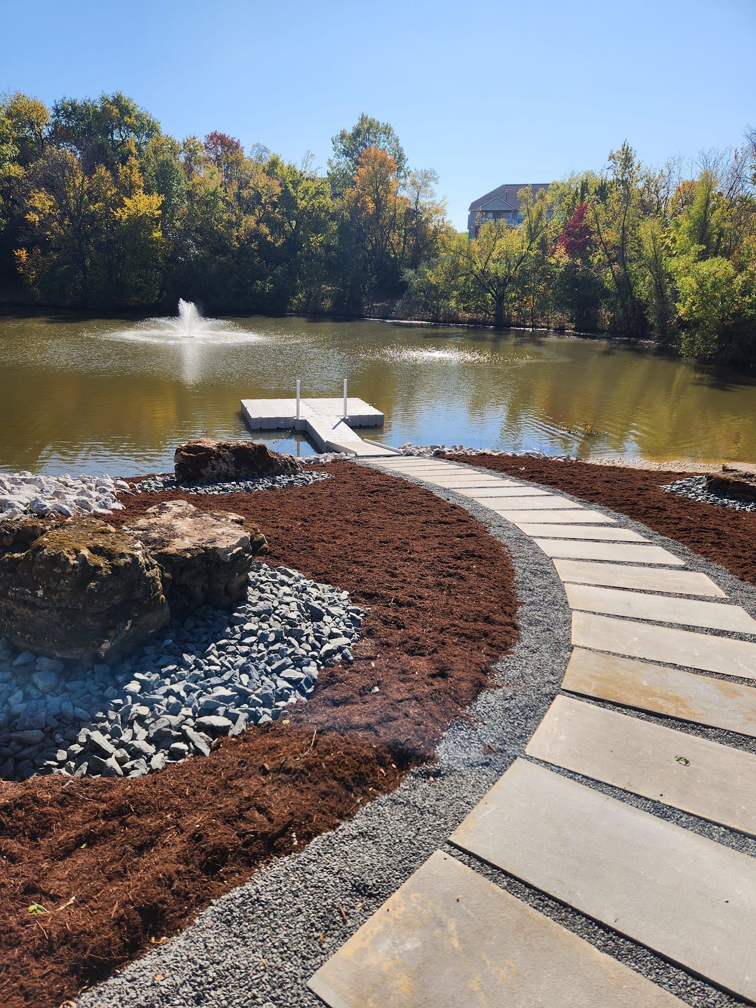 Stone path curves towards a pond with a dock and fountain. Foliage and trees surround the water on a sunny day.