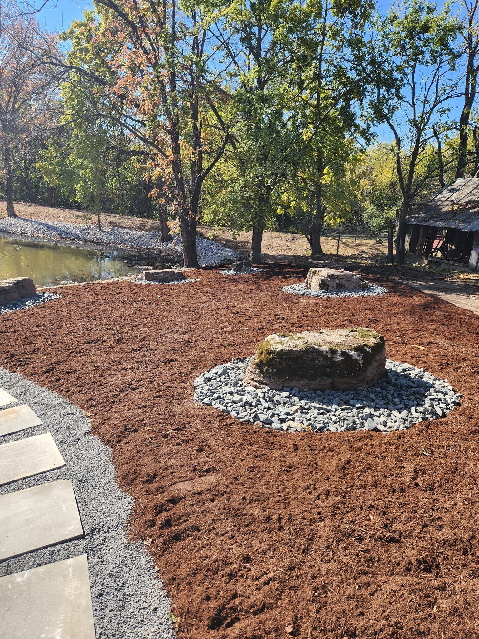 A stone rock in the center of a brown mulch bed surrounded by stones, a pond, and trees. A paved path leads to the scene.