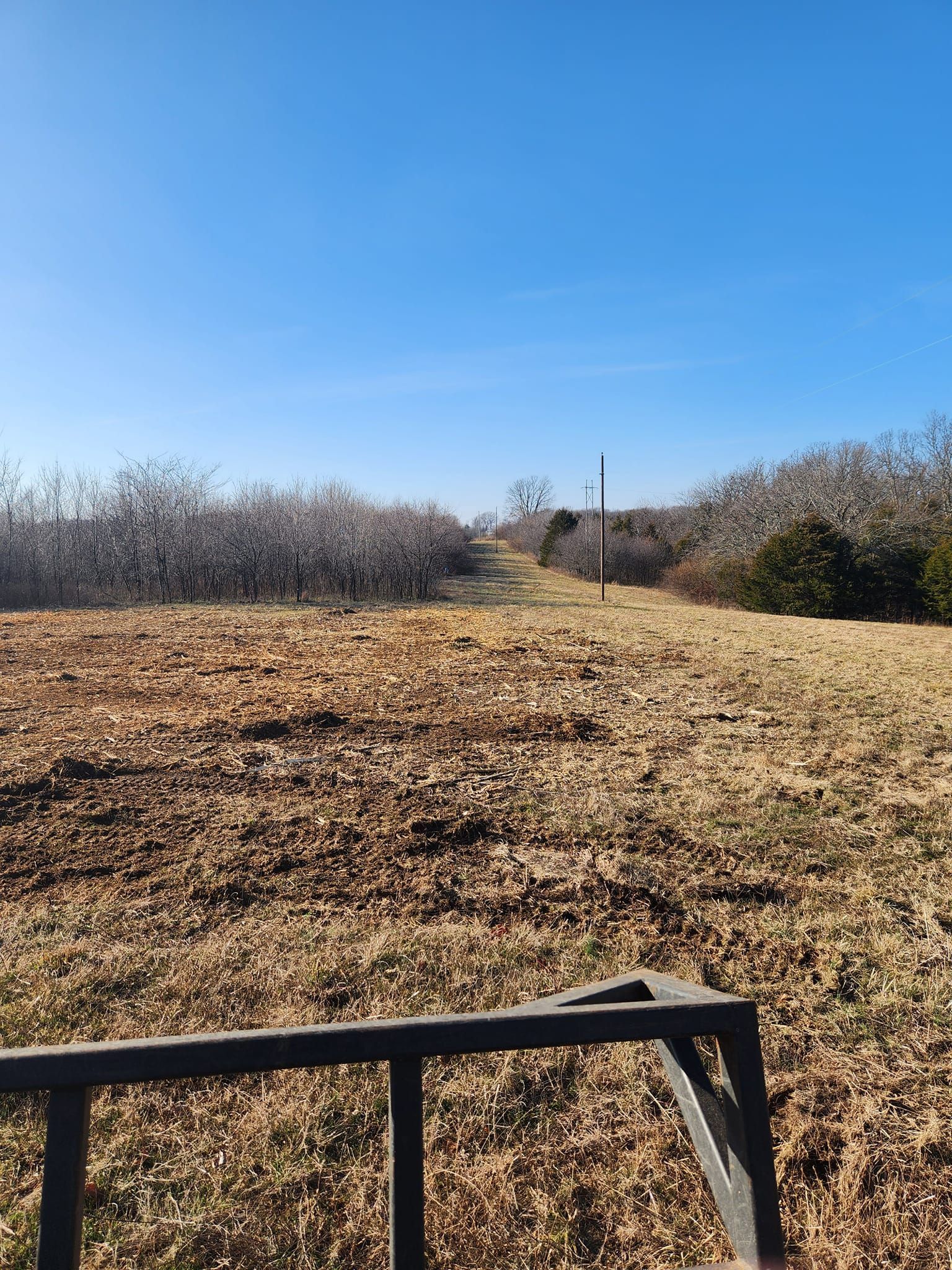 A path through dry grass and brush leads to a distant fence post under a clear blue sky.