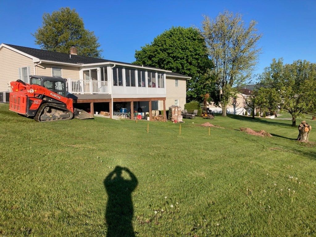 A red skid steer parked near a house with a deck; a person's shadow is in the foreground. A grassy yard slopes downhill.
