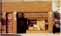 Small storefront with a red door, large front window, and brick facade on a sunny street.