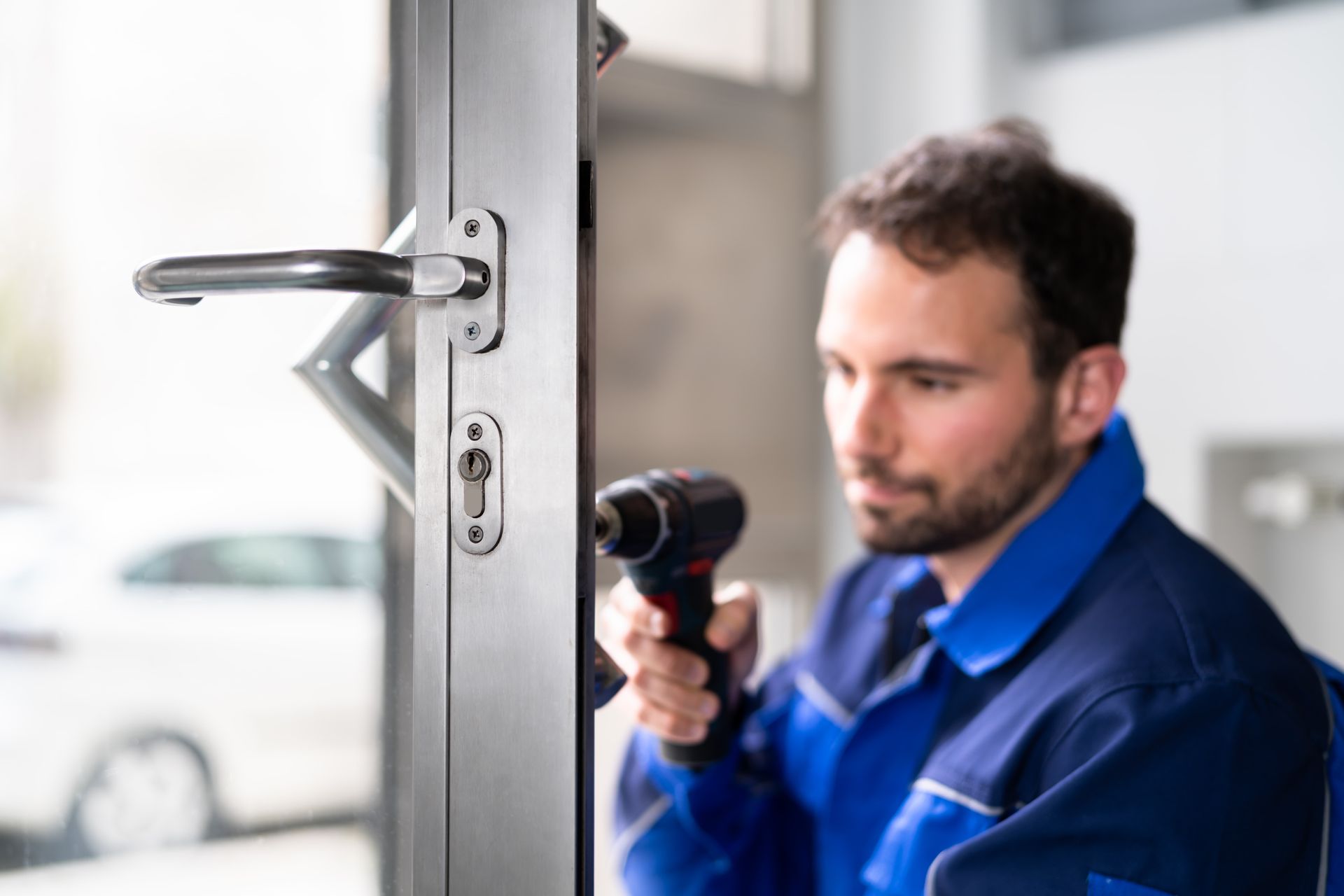 Worker in blue coveralls using a power drill on a door lock by a glass door.