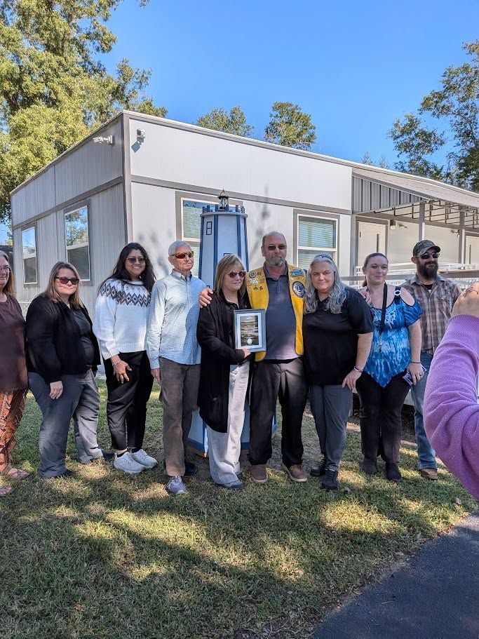 Group of people outside a white building, some holding a framed photo on a sunny day.
